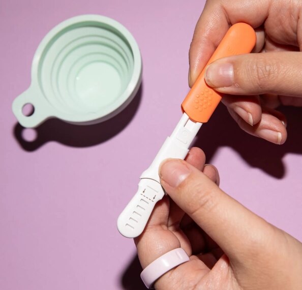 Тест на беременность в руке на фоне розового стола с мерной чашкой.