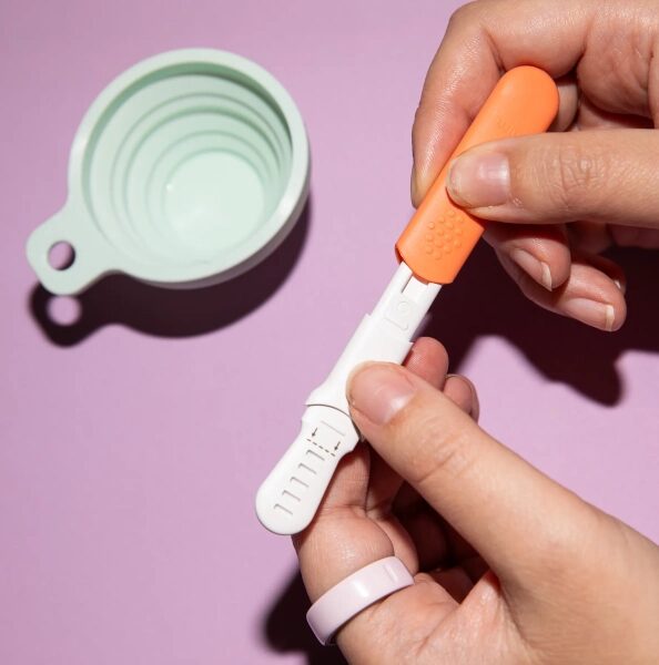 Тест на беременность в руке на фоне розового стола с мерной чашкой.