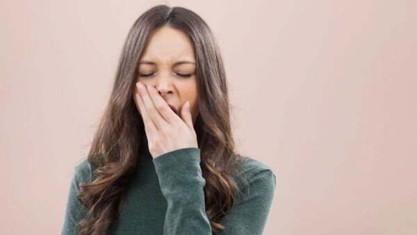 Женщина зевает на фоне бежевой стены, выражая усталость и сонливость.