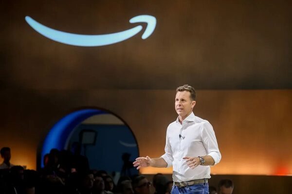 Человек в белой рубашке на сцене с логотипом Amazon на фоне.