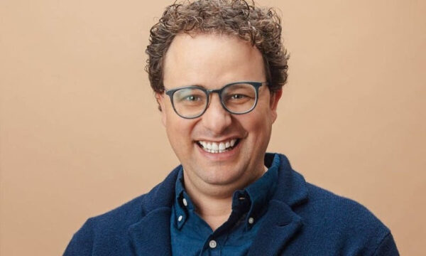 Улыбающийся мужчина в очках на бежевом фоне.