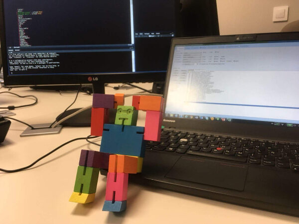 Многоцветный кубик-робот сидит на ноутбуке рядом с монитором, отображающим код.