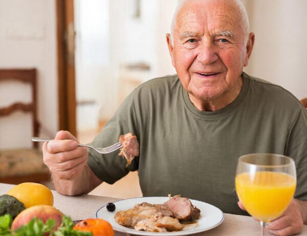 Мужчина в зеленой футболке ест мясо и пьет сок за столом дома, рядом свежие фрукты и овощи.