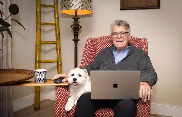 Мужчина за ноутбуком в кресле, рядом собака и кружка на столике.