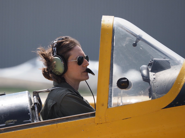 Кэти Мак сидит в кресле пилота желтого самолета. На ней зеленая куртка и наушники.. 