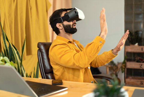 Человек в жёлтой рубашке использует VR-гарнитуру за столом с ноутбуком.