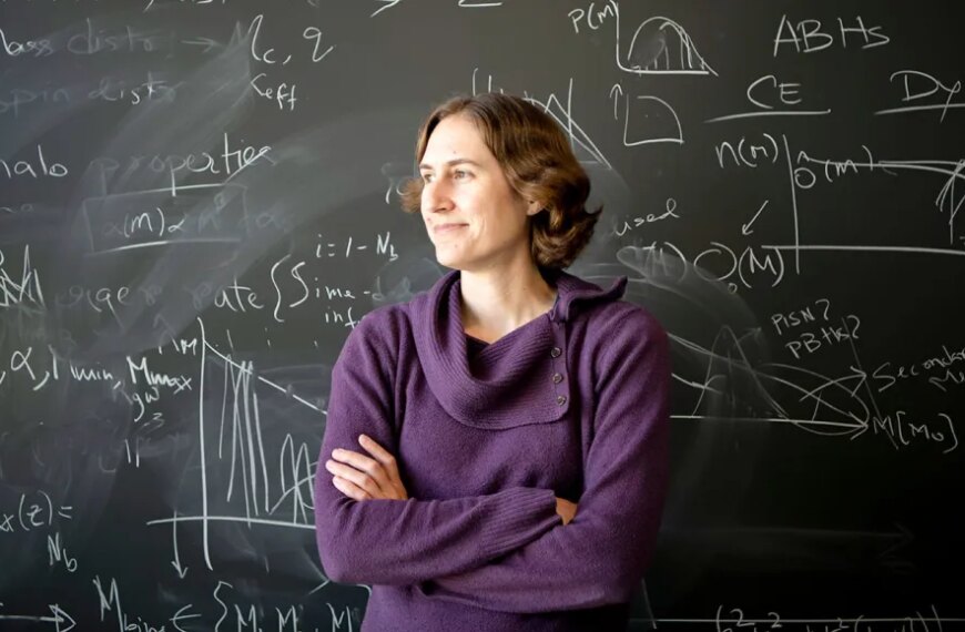 Женщина в фиолетовом свитере возле доски с формулами в классе.