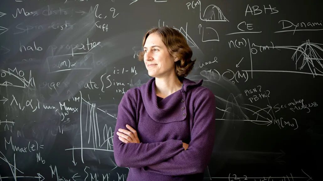 Женщина в фиолетовом свитере возле доски с формулами в классе.