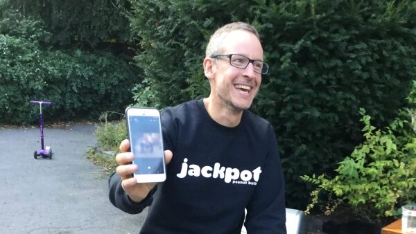 Человек в свитере с надписью "jackpot" показывает телефон на фоне зелени.