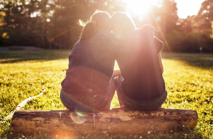 Две девушки сидят на бревне в лучах солнца на лужайке.