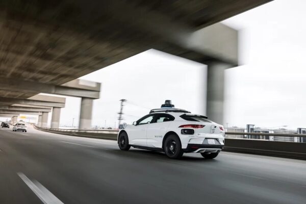 Белый автономный автомобиль едет по скоростной трассе под мостом.