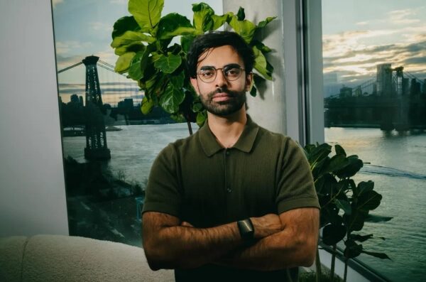 Человек в зелёной рубашке на фоне окна с видом на мост и реку в городской обстановке.