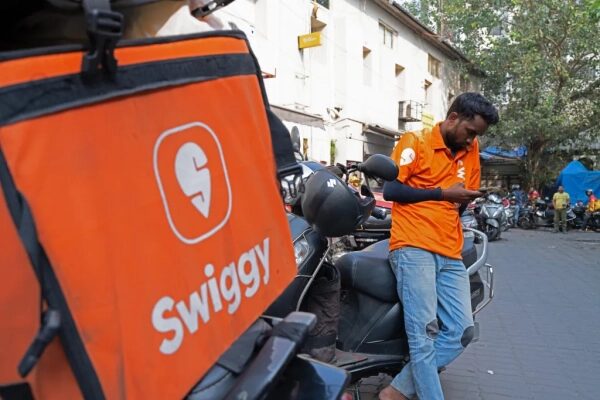 Курьер Swiggy на улице, ожидает заказ, опираясь на мотоцикл.