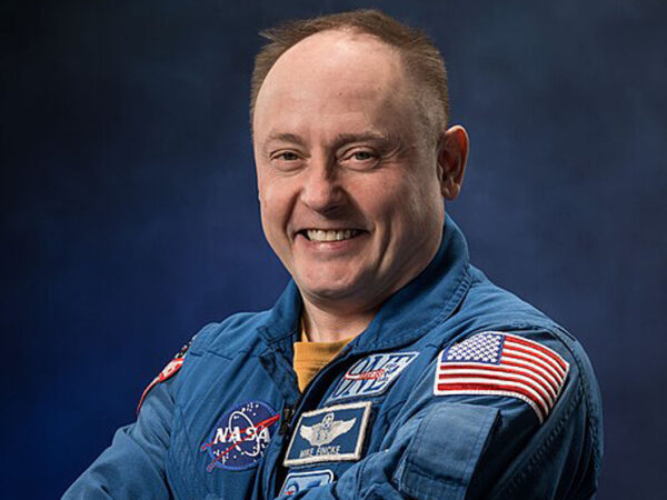 Астронавт в форменной одежде NASA с эмблемами и флагом США на рукаве.