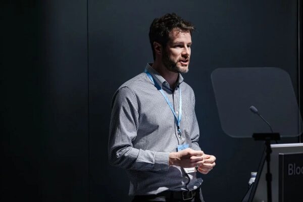 Человек в рубашке выступает на конференции с микрофоном и беджем.