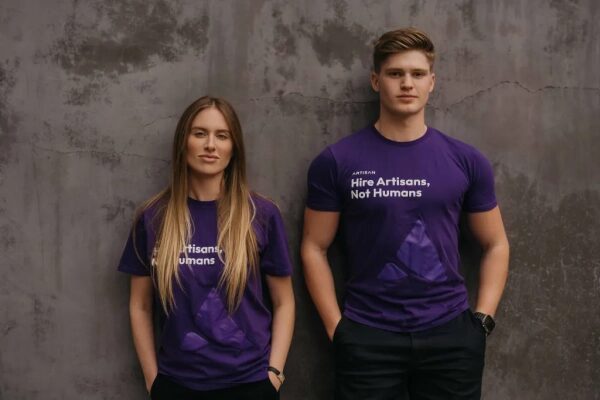 Два человека в фиолетовых футболках с надписью, стоят перед серой стеной.