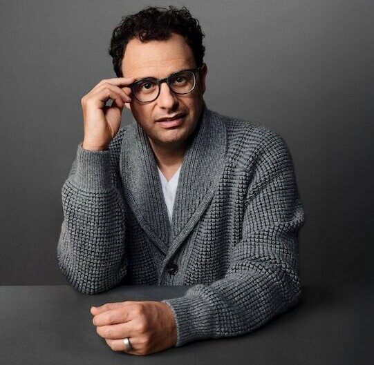 Мужчина в сером вязаном кардигане сидит за серым столом.