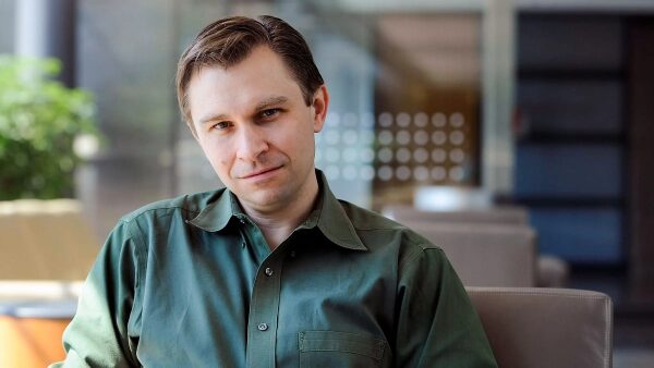 Мужчина в зеленой рубашке сидит в современной обстановке, смотрит в камеру.