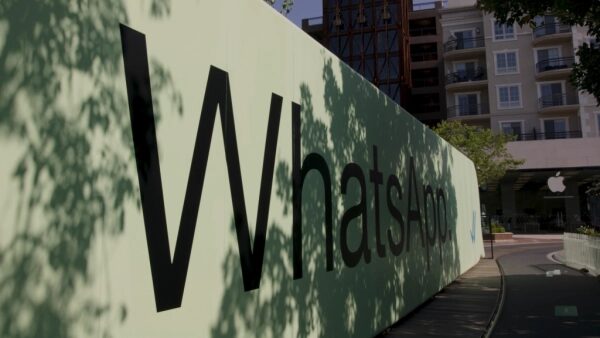 Стена с логотипом WhatsApp на улице в тени деревьев, виден магазин Apple.