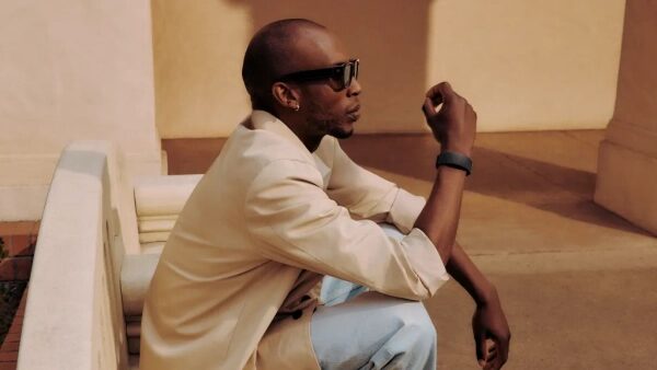 Человек в солнцезащитных очках и стильной одежде сидит на улице, задумчиво смотрит вдаль.