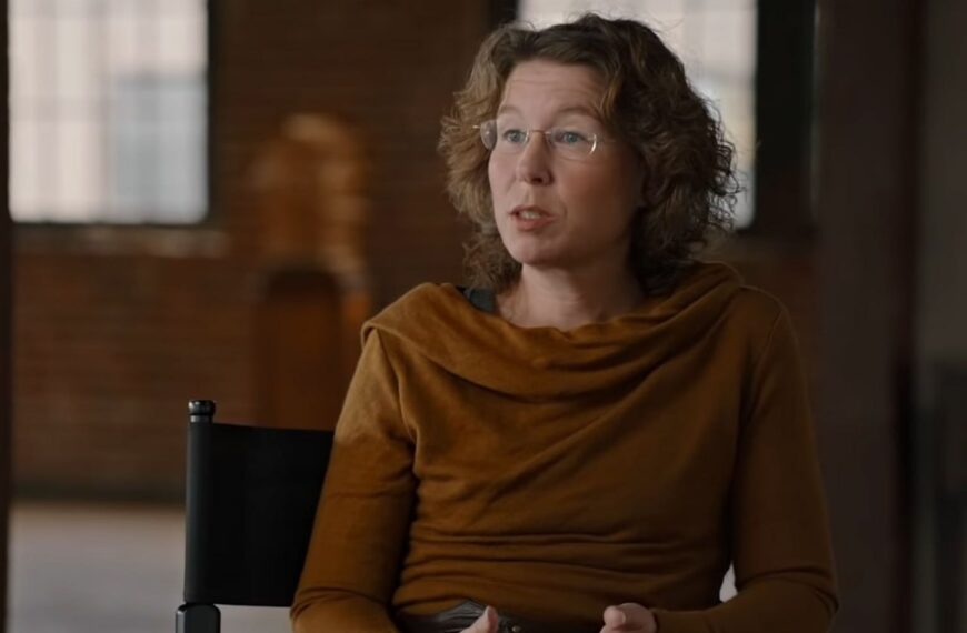 Человек в коричневой одежде сидит на черном стуле в помещении с кирпичными стенами.