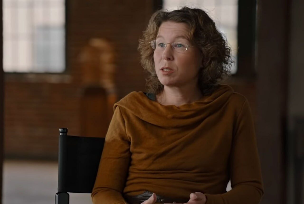 Человек в коричневой одежде сидит на черном стуле в помещении с кирпичными стенами.