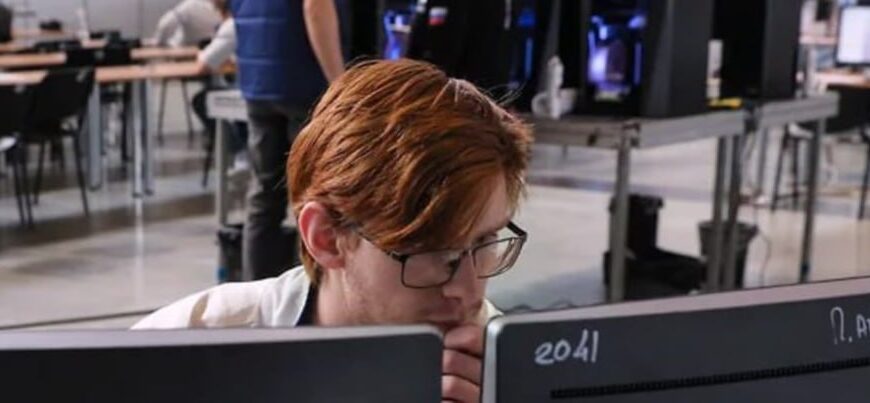 Человек с рыжими волосами в очках работает за компьютером в офисе с 3D-принтерами.