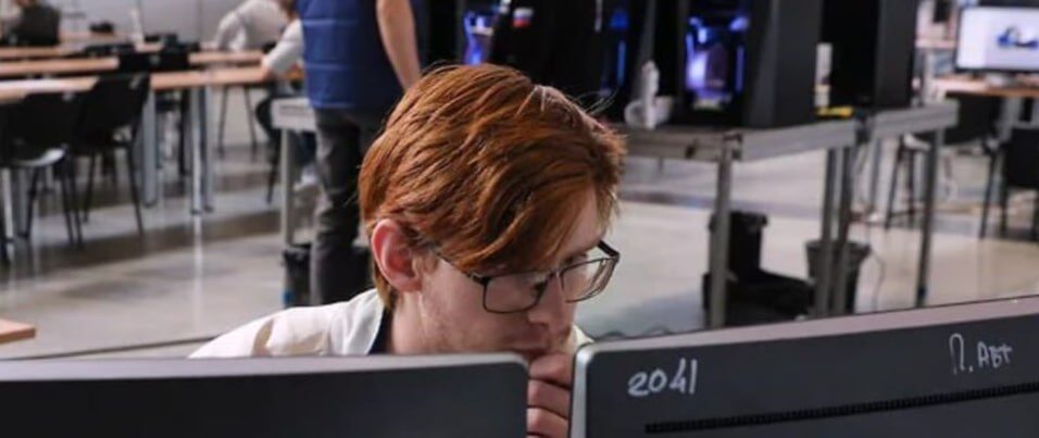 Гос-IT снова с льготами — но не для всех Минцифры... 2 Человек с рыжими волосами в очках работает за компьютером в офисе с 3D-принтерами.