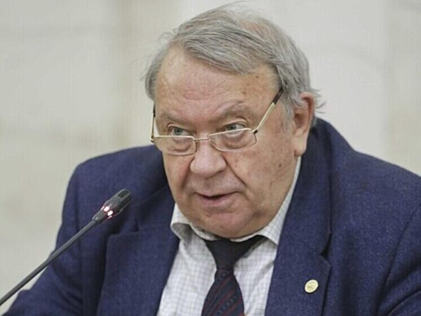 Мужчина в костюме и галстуке говорит в микрофон, белый фон.