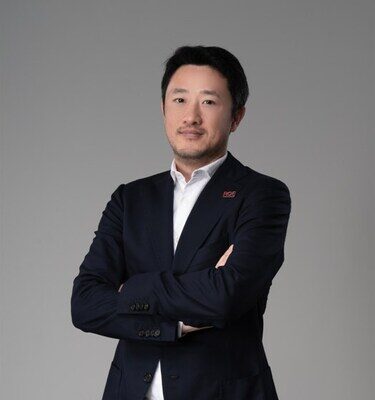 Мужчина в темном костюме на сером фоне, скрестивший руки на груди.