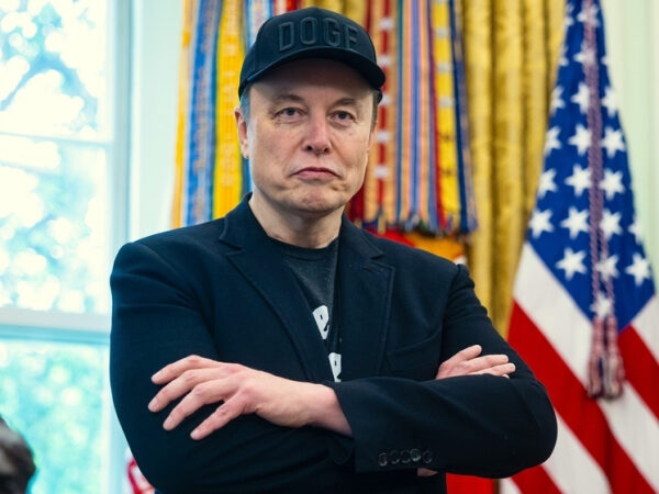 Человек в черной куртке и кепке "DOGE" на фоне американского флага.