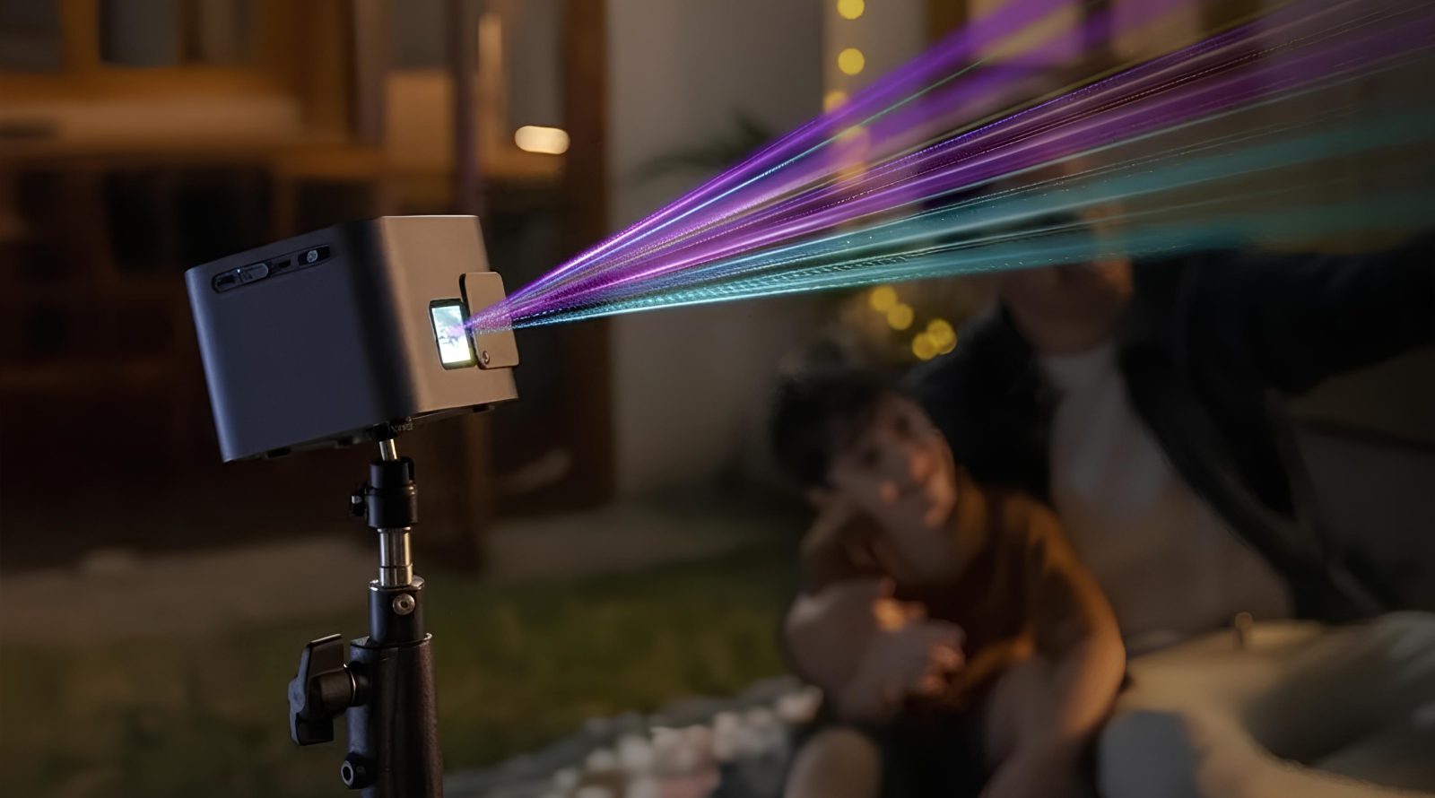 Рисование светом: лазерный проектор Laserabc A1 3 Проектор с цветными лучами в ночном саду, на фоне размытые фигуры людей.