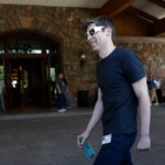 Мужчина в черной футболке и с бутылкой идет у входа в здание с каменными стенами.