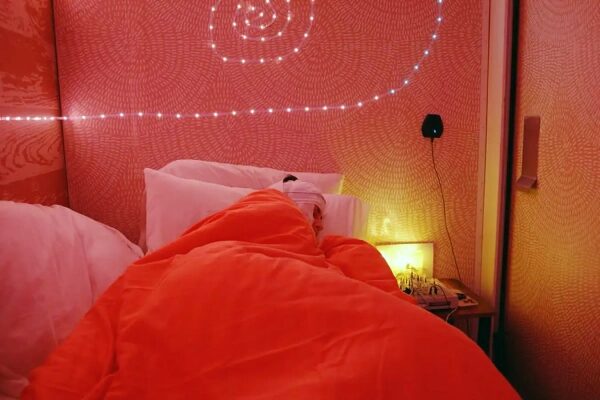 Человек спит в кровати с оранжевым одеялом и светодиодной подсветкой на стене.