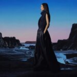 Женщина в длинном платье позирует на фоне скалистой местности на закате.