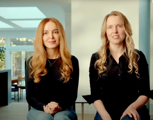 Две женщины с длинными волосами сидят в светлой комнате, одеты в черное.