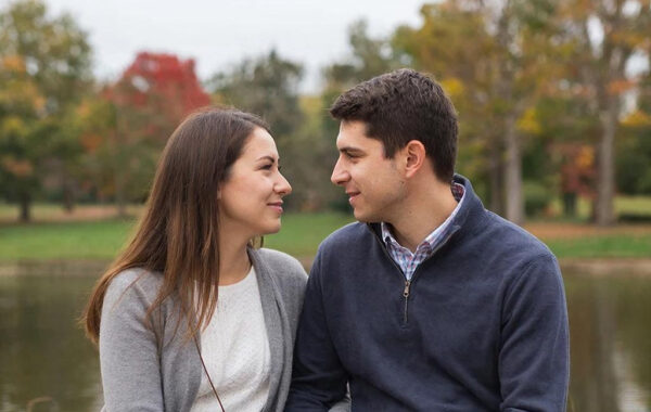 Пара на природе осенью у пруда, деревья с разноцветной листвой на заднем плане.