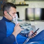 Мужчина с ингалятором в руках, сидит на диване и использует планшет.