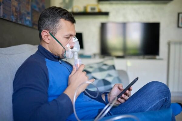Мужчина с ингалятором в руках, сидит на диване и использует планшет.