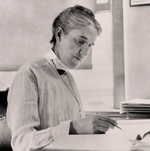 Черно-белое фото Генриетты Свон Ливитт. Она сидит за своим столом и пишет в блокноте.