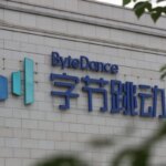 Логотип ByteDance на кирпичной стене, частично скрыт ветвями деревьев.