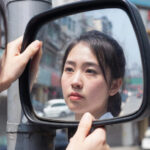 Зеркало на улице, женщина смотрит, держась за края, отражение городского пейзажа.