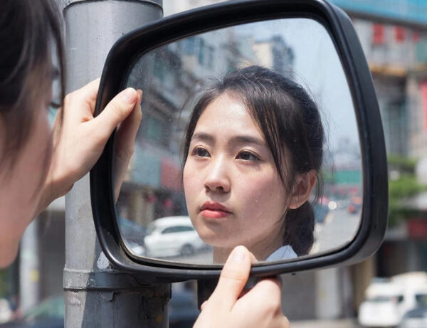 Зеркало на улице, женщина смотрит, держась за края, отражение городского пейзажа.