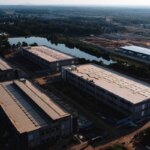 Промзона с крупными складами у озера на фоне зелёного пейзажа, вид сверху.