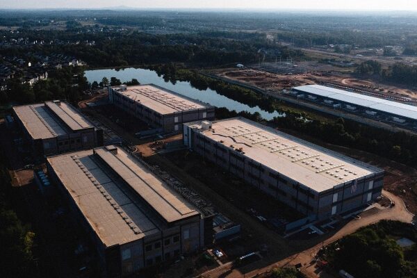 Промзона с крупными складами у озера на фоне зелёного пейзажа, вид сверху.