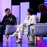 Три человека обсуждают на конференции, сидя на серых креслах.