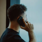 Мужчина в черной футболке разговаривает по телефону у окна.