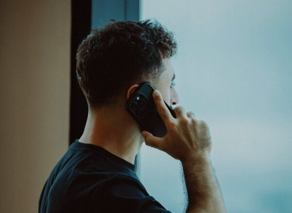 Мужчина в черной футболке разговаривает по телефону у окна.