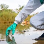 Человек в защитной одежде берет пробы воды из озера для анализа загрязнений.