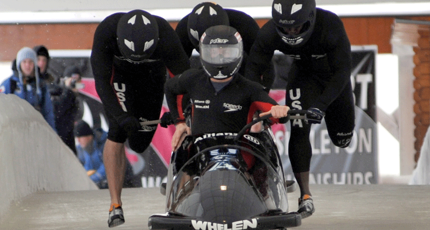 Спортсмены в бобслее на старте, команда в черной униформе с буквами USA.